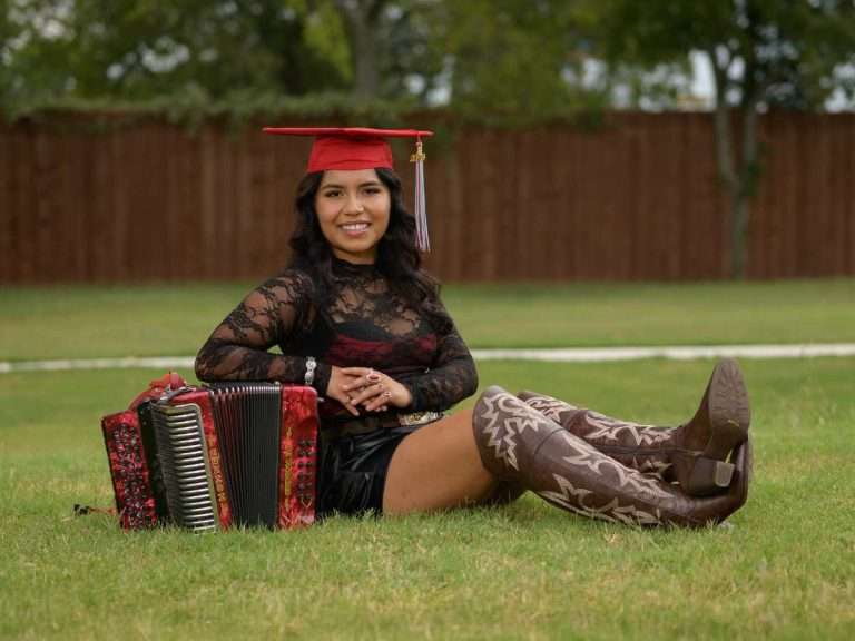 Skyline High School senior graduate in cap and gown sitting in the grass holding her musical instrument during a DFW senior portrait session